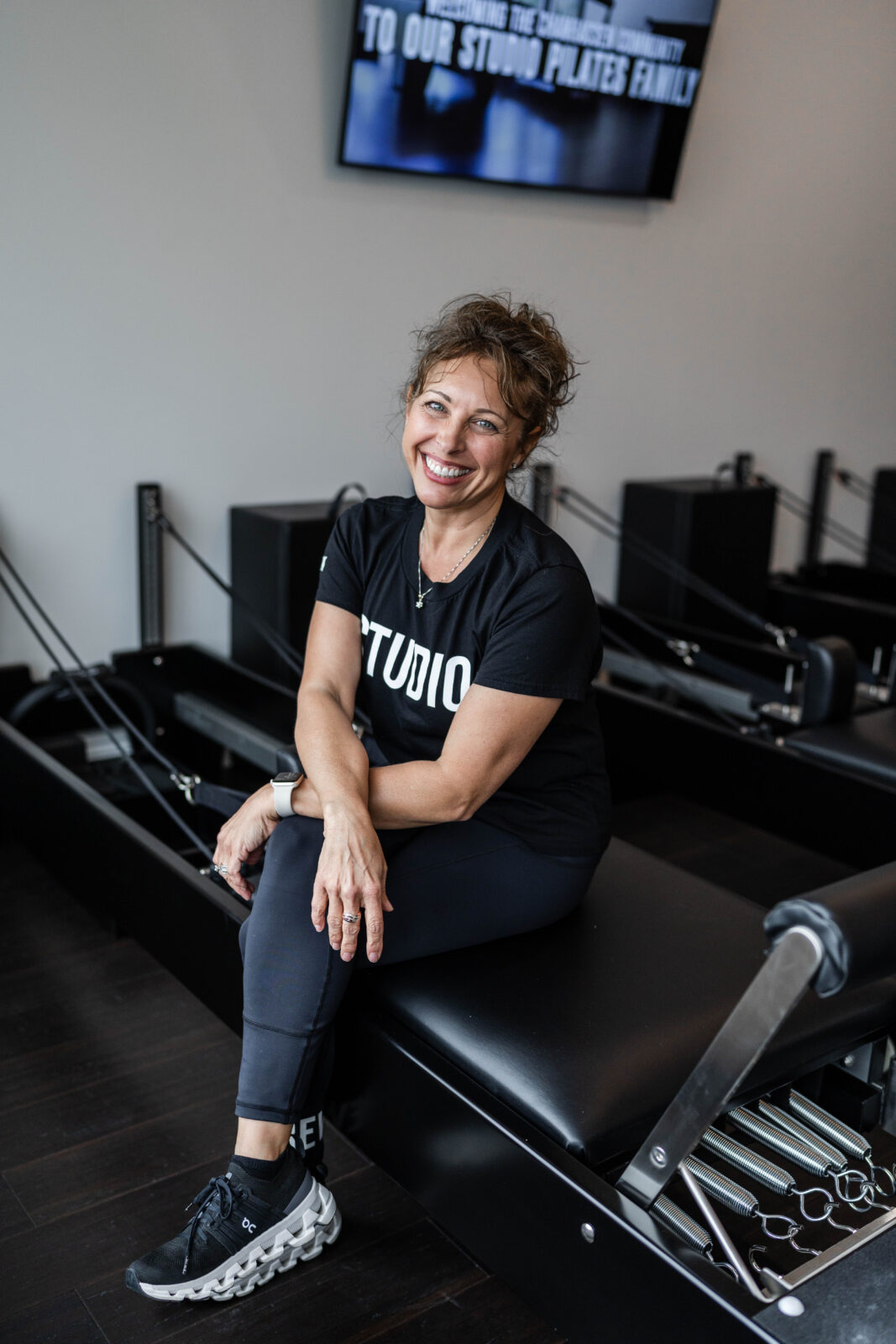 Woman wearing a black t-shirt with white text sitting in a Pilates studio with reformer machines.