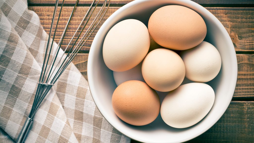 Bowl with six eggs of varied colors on wooden surface beside a metal whisk and checkered cloth.