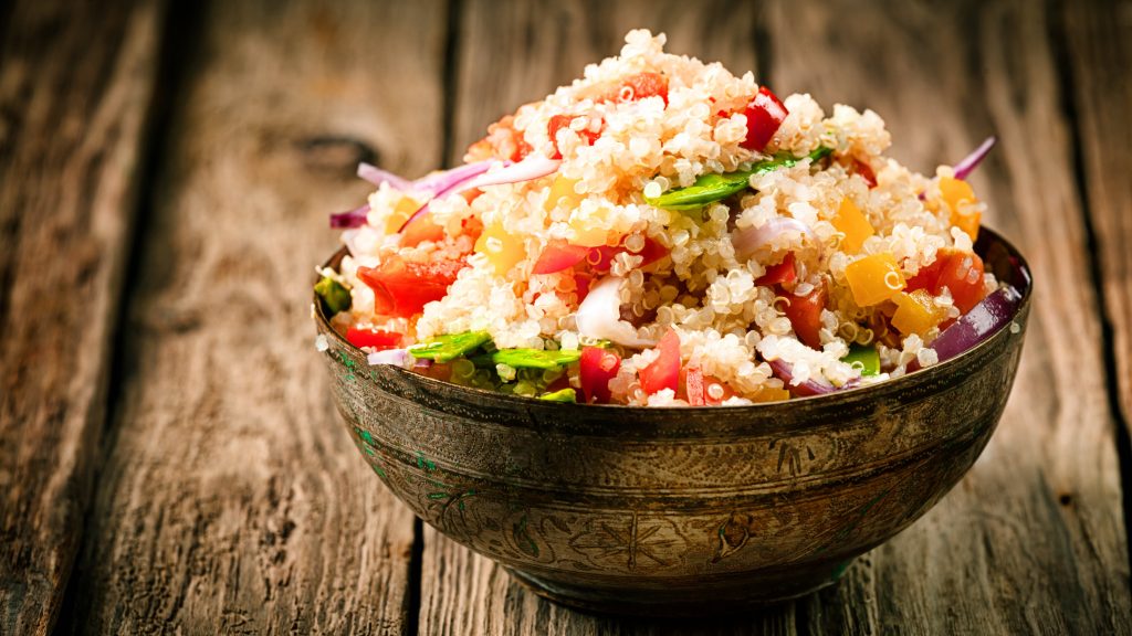 Bowl of quinoa salad with diced tomatoes, red onions, green vegetables, and yellow bell peppers.
