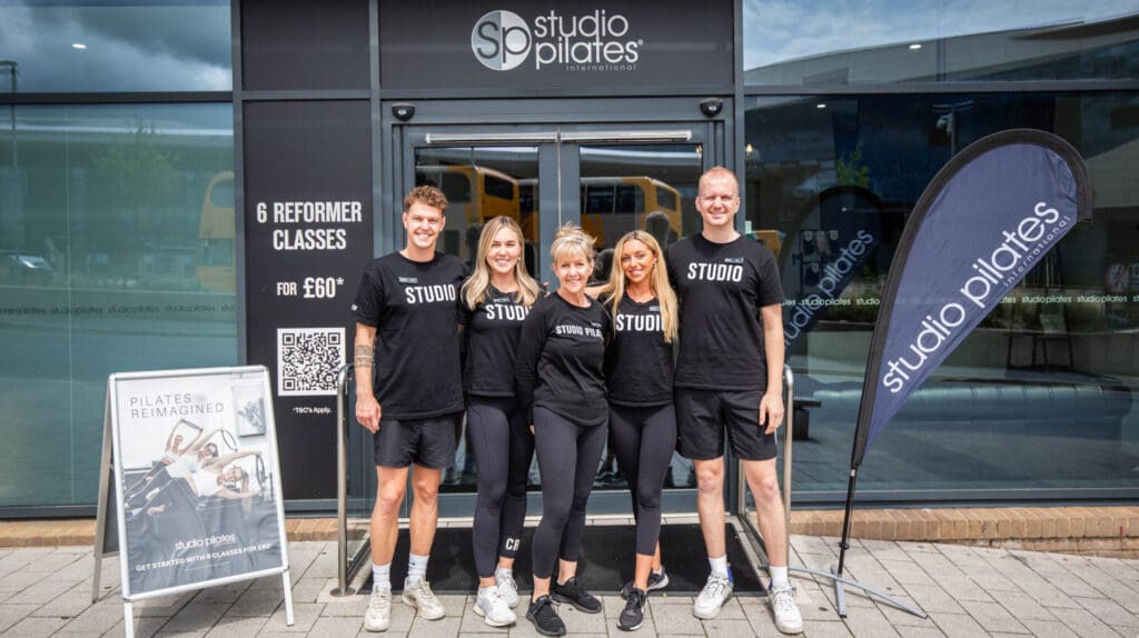 Five Studio Pilates staff stand outside the studio with promotional signs and a branded flag.