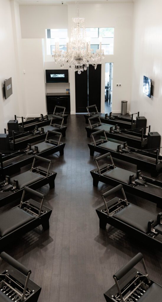 Pilates studio with two rows of black reformer machines, white walls, chandelier, and screens.