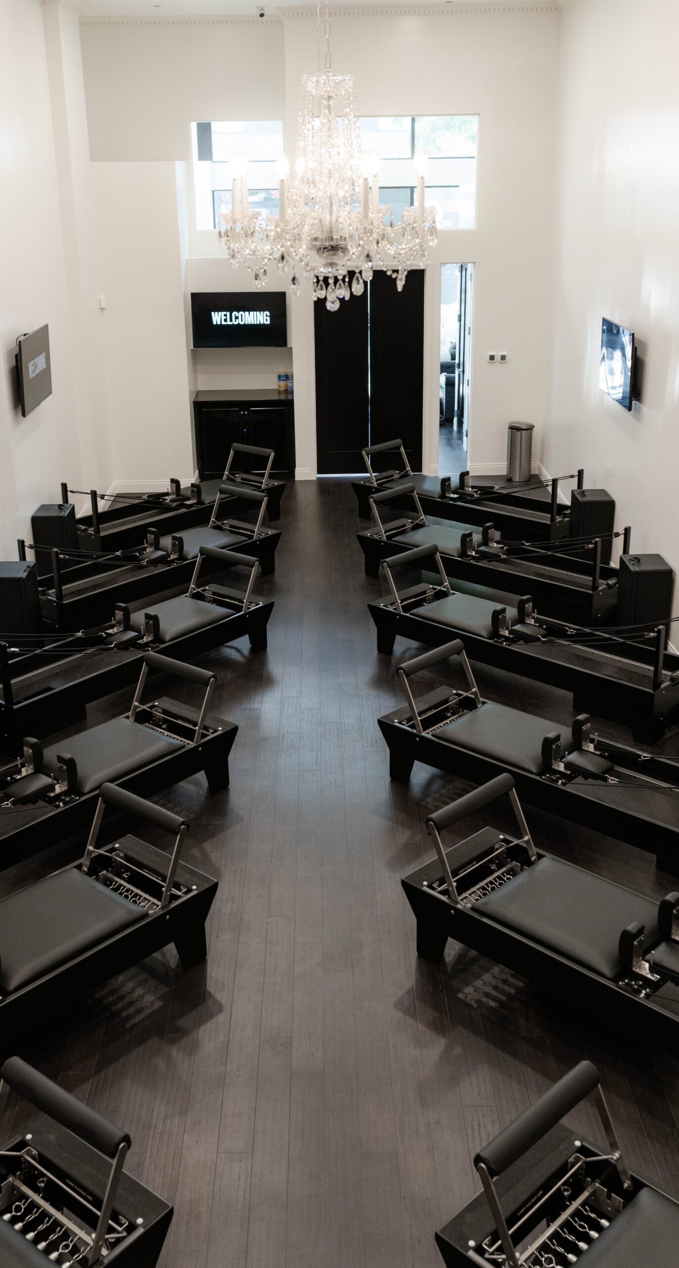 Pilates studio with two rows of black reformer machines, white walls, chandelier, and screens.