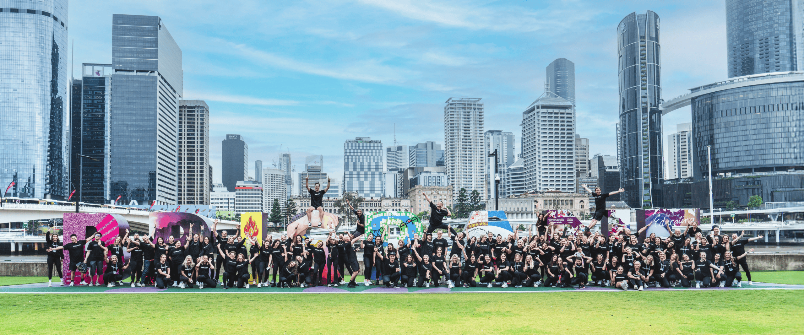 Large group in black shirts posing outdoors by colorful letters with city skyline background.